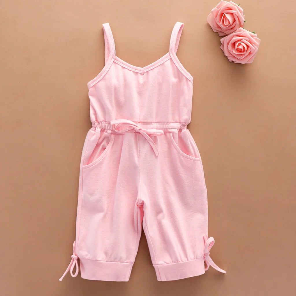 A pink solid color sling jumpsuit for babies, with a functional pocket and adjustable suspender straps, displayed against a neutral background with a few pink roses to the side.